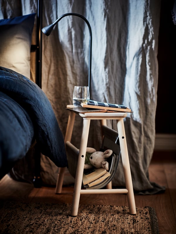 Un letto con biancheria blu scuro. Accanto, sopra a un tappeto, uno sgabello usato come comodino con sopra libri e un bicchiere - IKEA