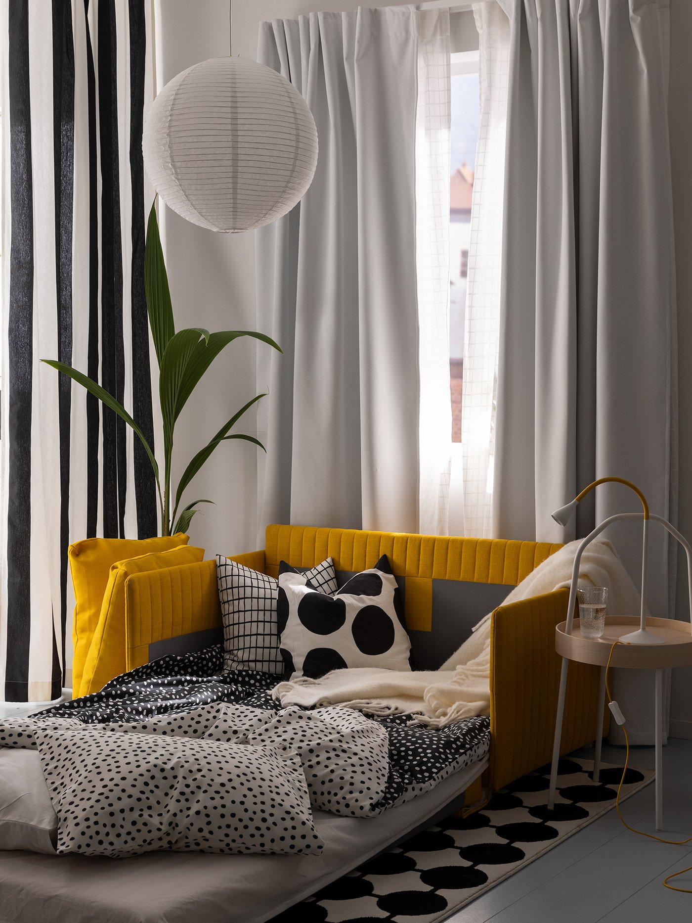 A yellow FRIDHULT sofa-bed, shown as a bed, stands at a window with KORGMOTT block-out curtains and GINSTMOTT curtains.