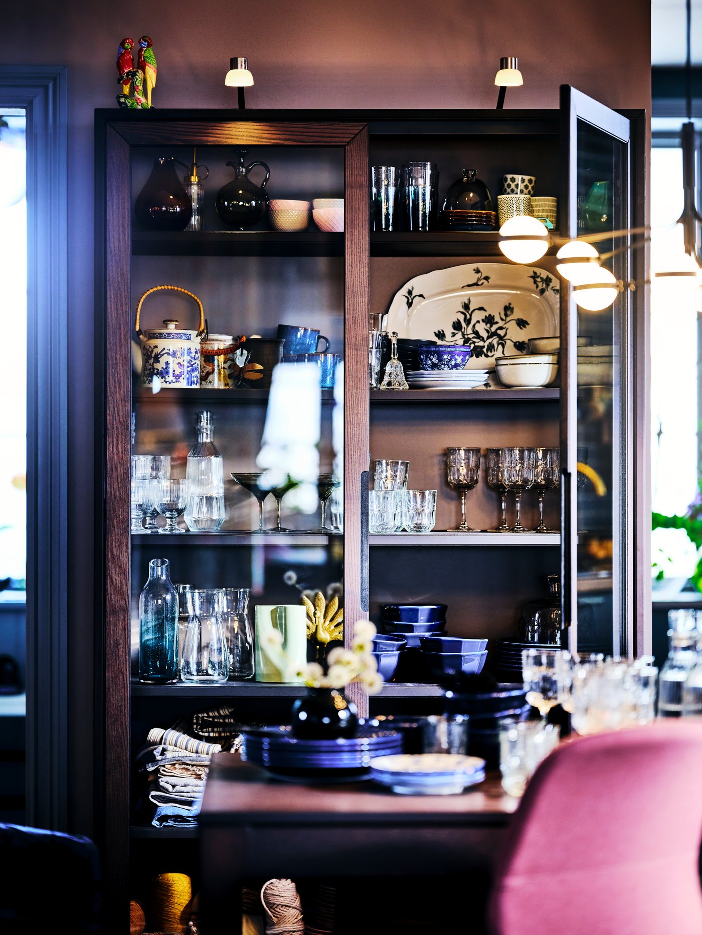 A brown REGISSÖR glass-door cabinet in a dining area with one door ajar housing a collection of crockery and glassware.