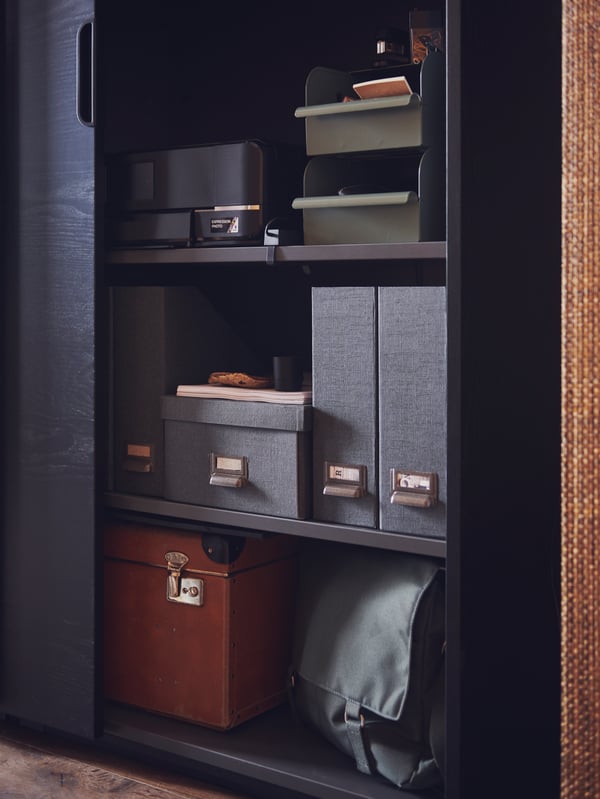A black GALANT cabinet with the sliding door open showing three shelves holding magazine files, a tray and storage boxes.