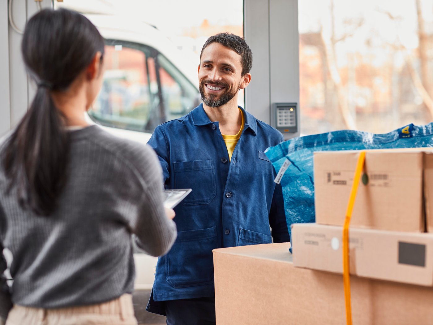A co-worker in a blue shirt is delivering some parcels to a woman in the foreground with a grey sweater.