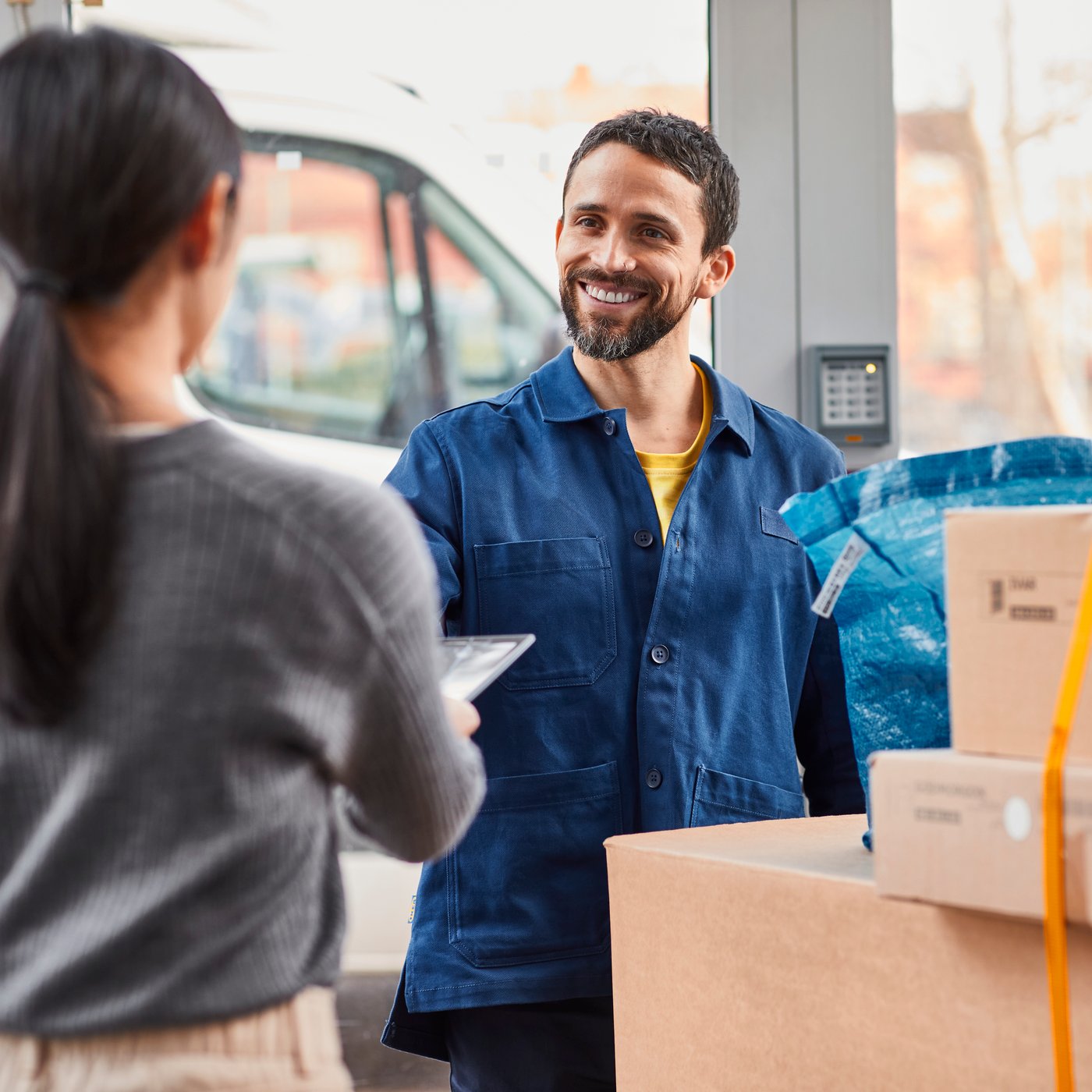 A co-worker in a blue shirt is delivering some parcels to a woman in the foreground in a grey sweater.