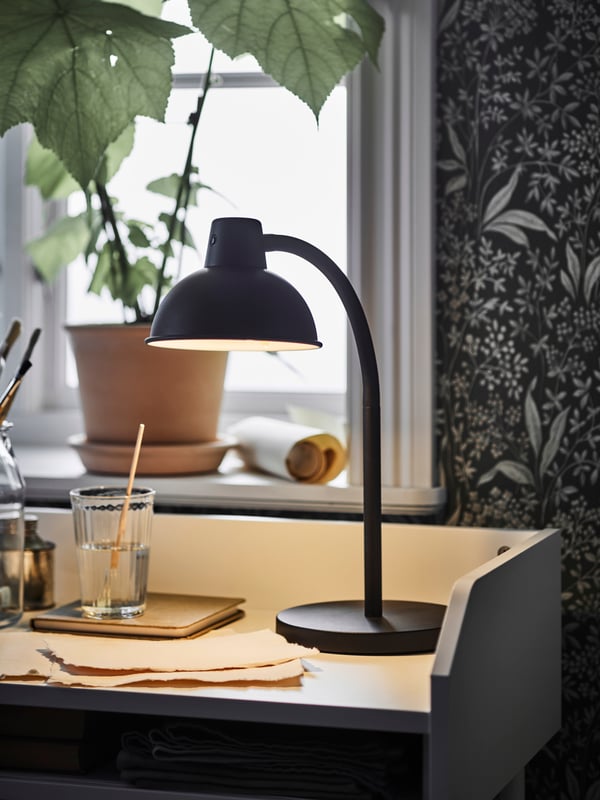 A black SKURUP work lamp stands near a VARDAGEN glass in a corner of the top of a white HAUGA desk near a window.