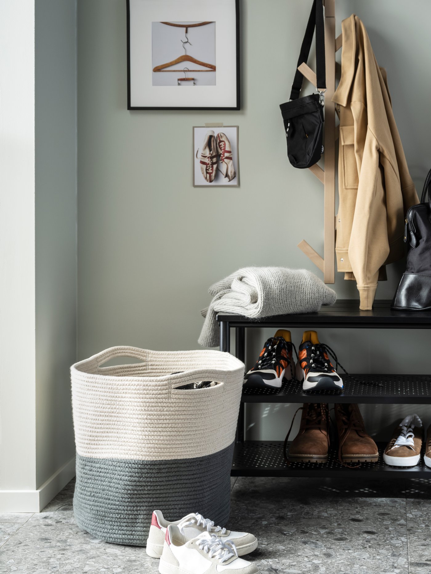 Part of a hallway with a two-coloured TJABBIG basket placed on the floor next to a black PINNIG bench with shoe storage.
