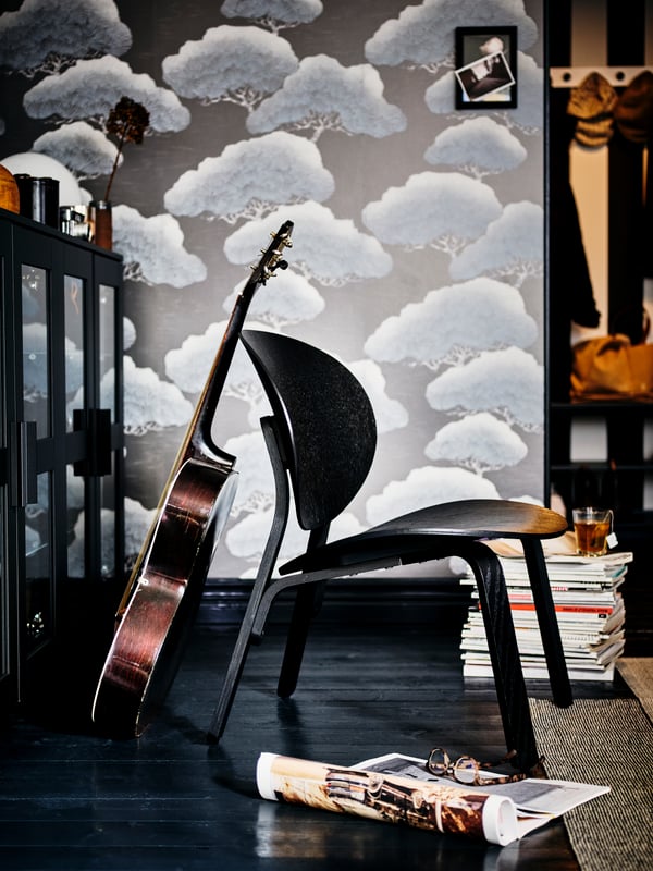 Le détail d’une pièce avec du papier peint à motif, le sol en bois foncé et une guitare qui s’appuie sur l'arrière d’une chaise noire.