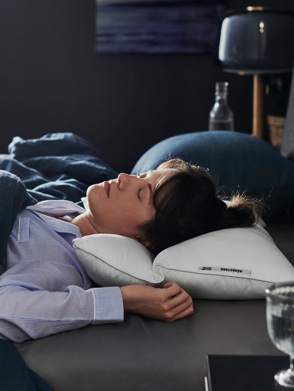 A woman wearing blue pyjamas sleeps on her back in a large bed with her head on a NÄBBSTARR ergonomic pillow.