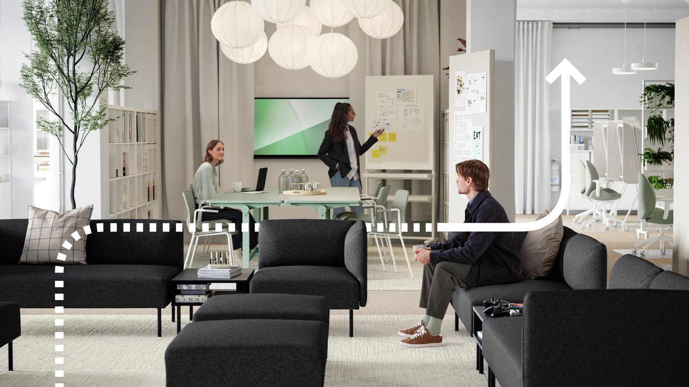 Ein junger Mann sitzt in einem Loungebereich, der aus mehreren dunkelgrauen LILLEHEM Sofamodulen und TUNSTA Tischen besteht, dahinter befindet sich ein Besprechungsbereich mit Whiteboard. 