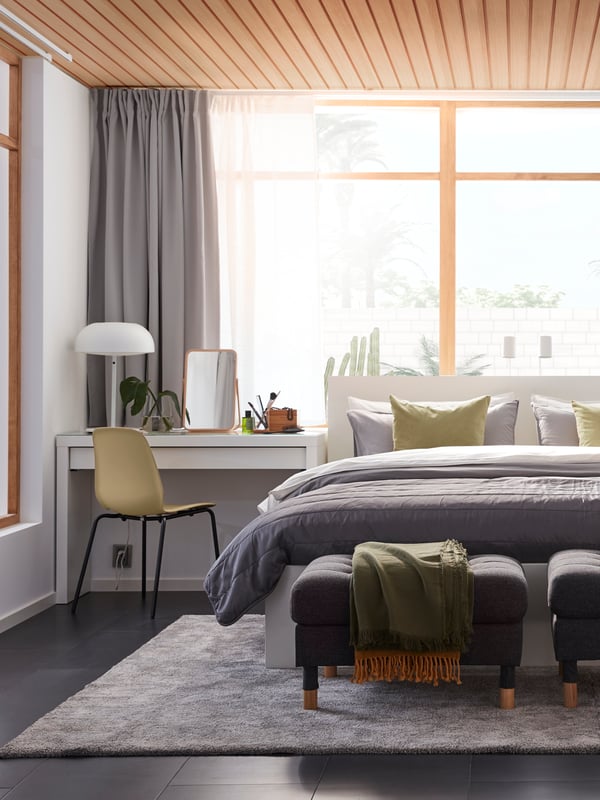 Part of a bedroom with a white bed and dressing table from the MALM series, a MORABO footstool and a large grey STOENSE rug.