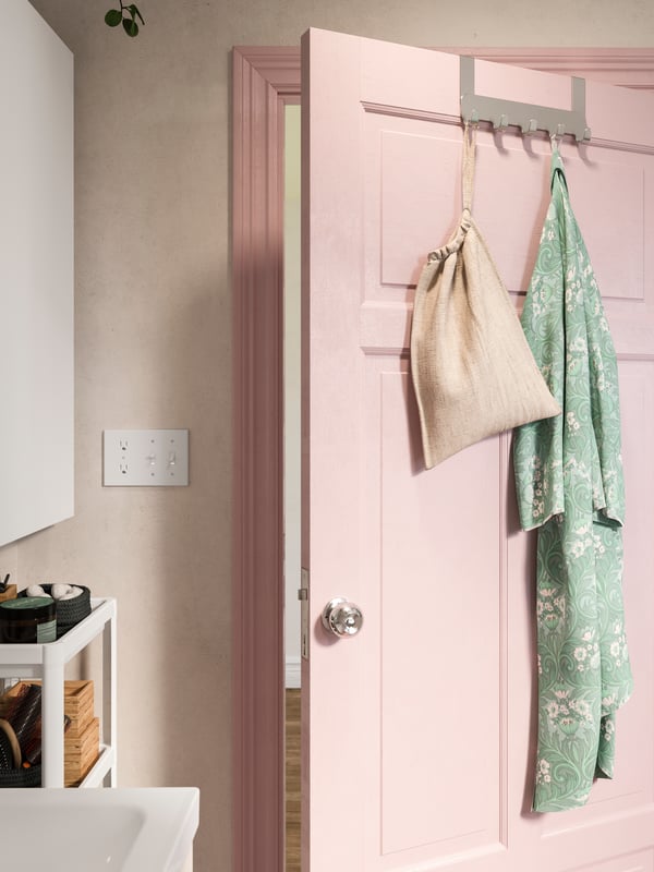BROGRUND hanger for door in stainless steel hangs on a pink bathroom door. A bag and a bathrobe hang on the hooks.