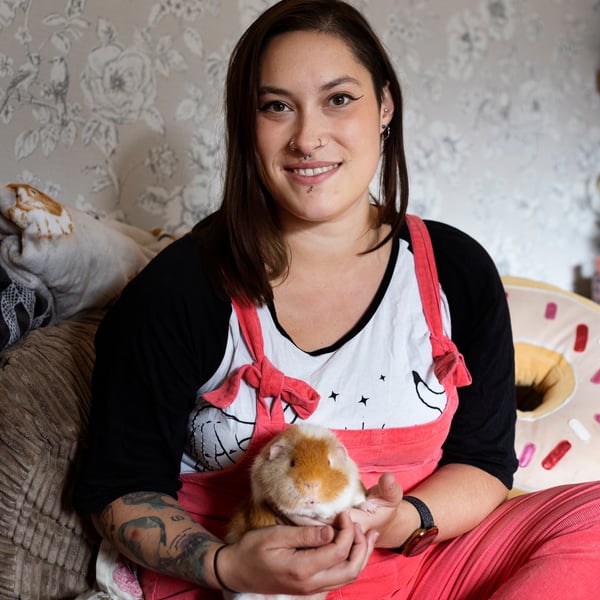 Happy woman sits on her bed smiling and looking at camera, while holding a small guinea pig.