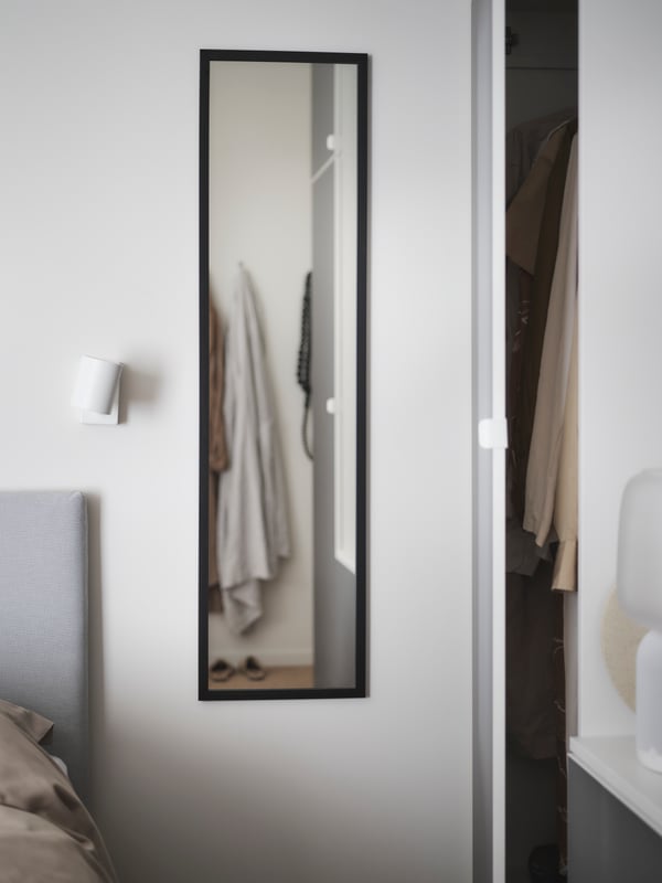 In a bedroom are a narrow and tall black LILJETRÄD mirror mounted on the wall, a white lamp, a white wardrobe and a grey bed.