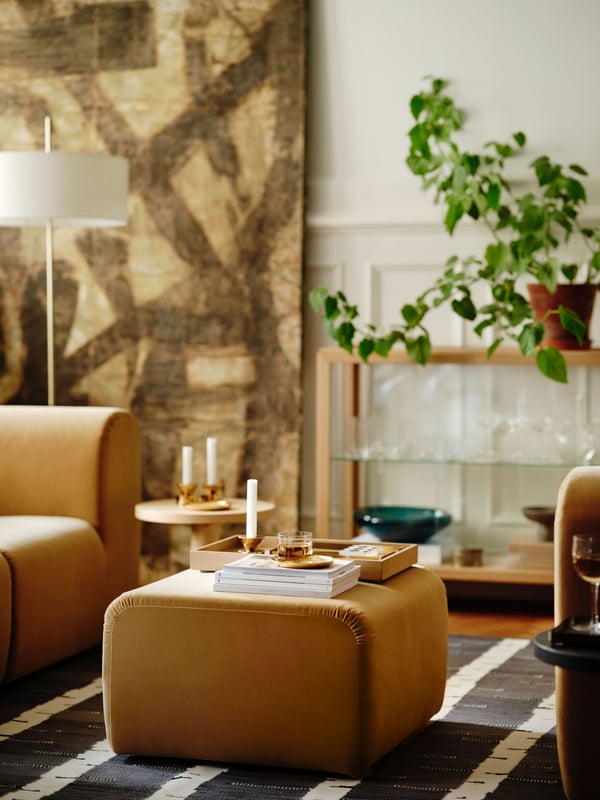 A beige STOCKHOLM 2025 pouffe with books, an oak veneer tray, a brass dish and candlestick/tealight holder, and more on top.