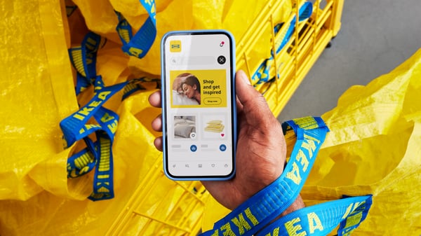 Smartphone mit geöffneter IKEA App vor gelben IKEA Einkaufstaschen mit blauen Griffen – zeigt Nutzerprofil, Text und Navigationssymbole.