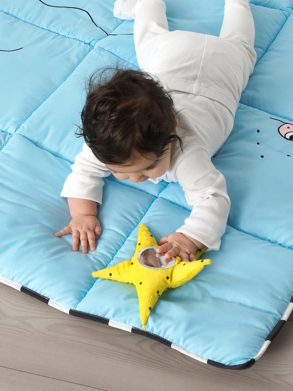 Bébé vêtu de blanc allongé sur le ventre sur un tapis de jeu KLAPPA bleu et tenant une étoile jaune avec un miroir.
