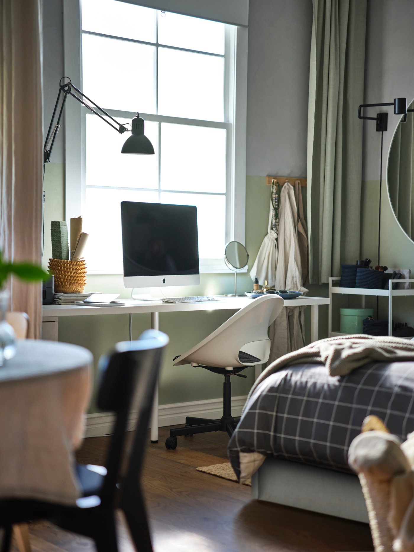 Un ordenador sobre un escritorio blanco LAGKAPTEN/ADILS con una silla giratoria LOBERGET/MALSKÄR con cojín, debajo de una ventana, junto a una cama.