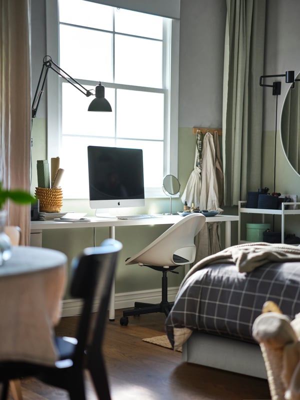 A computer atop a white LAGKAPTEN/ADILS desk with a LOBERGET/MALSKÄR swivel chair with pad, under a window, next to a bed.