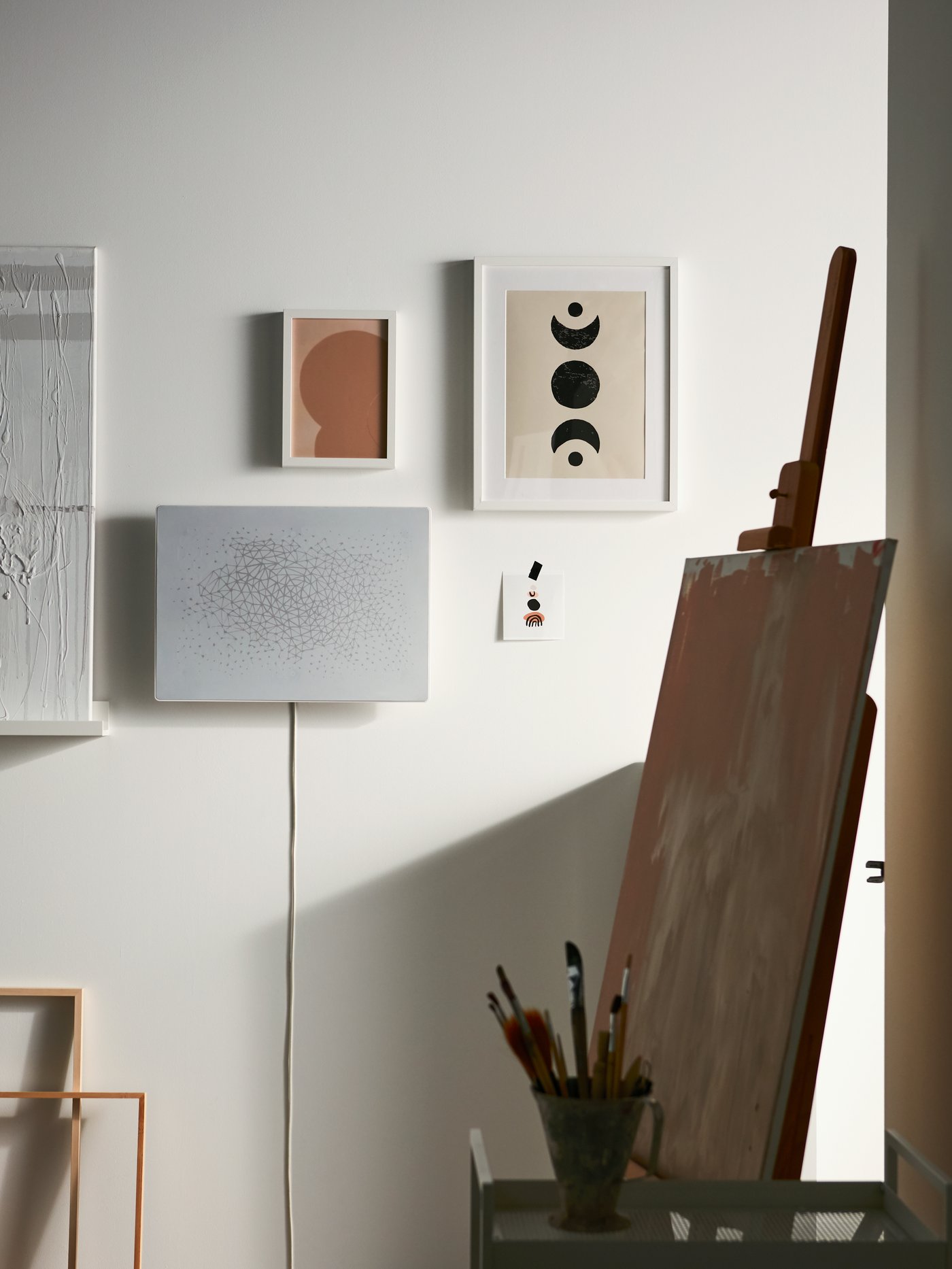 A white SYMFONISK picture frame with WiFi speaker is mounted on a wall with framed pictures and near a picture on an easel.