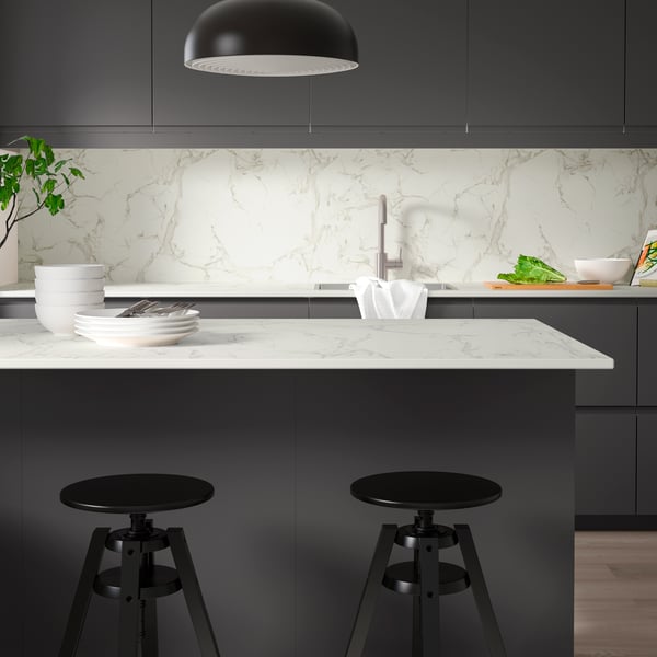 A kitchen with SKARARP custom-made worktop in white/grey on the kitchen island, tableware, and two black kitchen stools.
