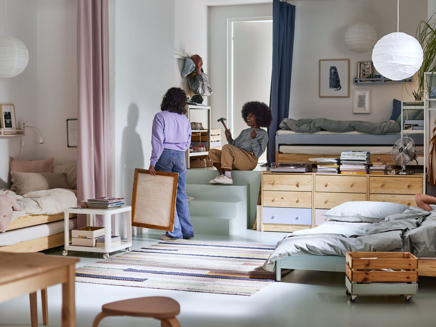Deux personnes décorant une pièce multifonctionnelle aux couleurs claires avec trois lits, des rangements, des tables d’appoint et un mini-escalier central.