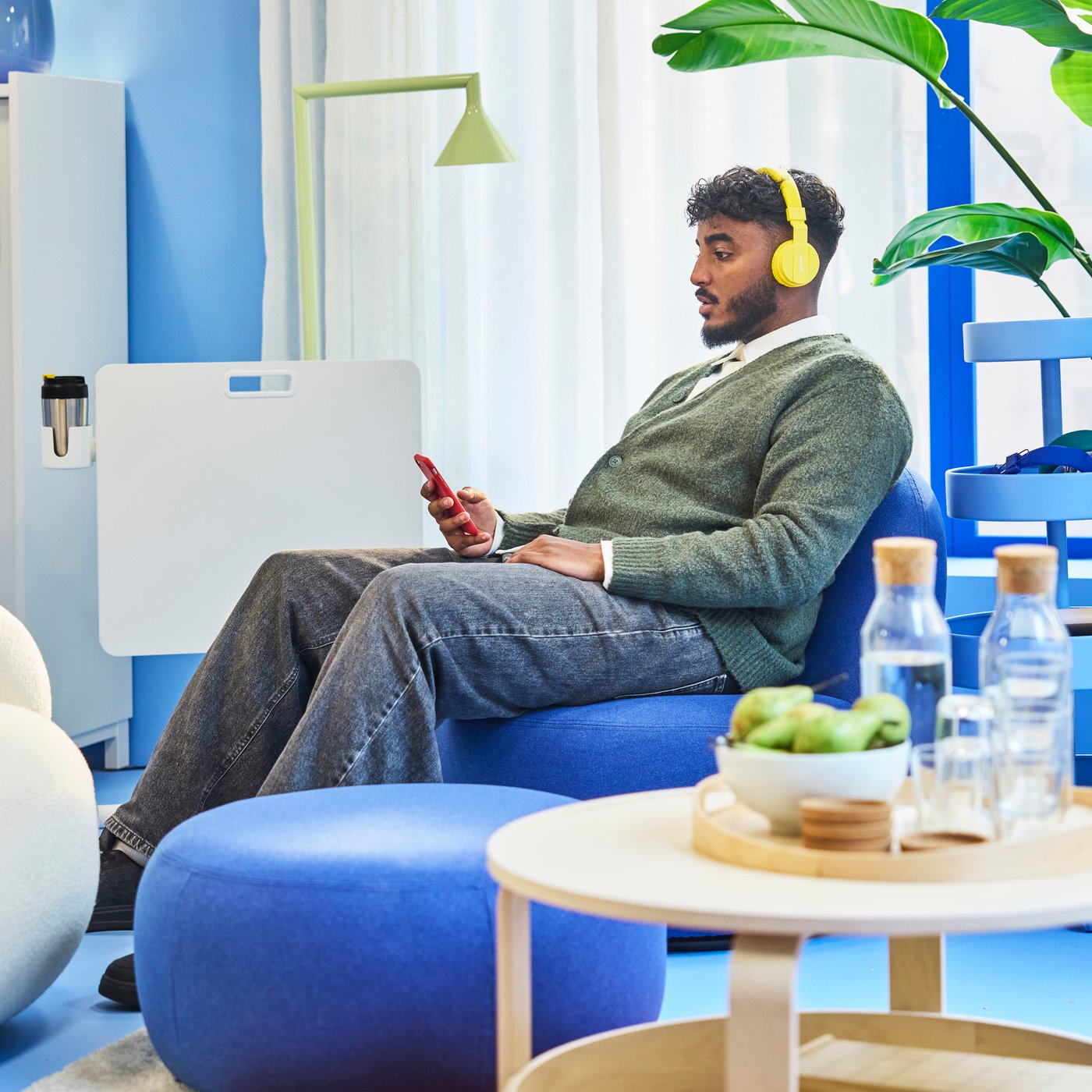 En person i grön tröja sitter i en blå stol med gula hörlurar och tittar på telefonen i ett ljust rum med växter och ett bord.