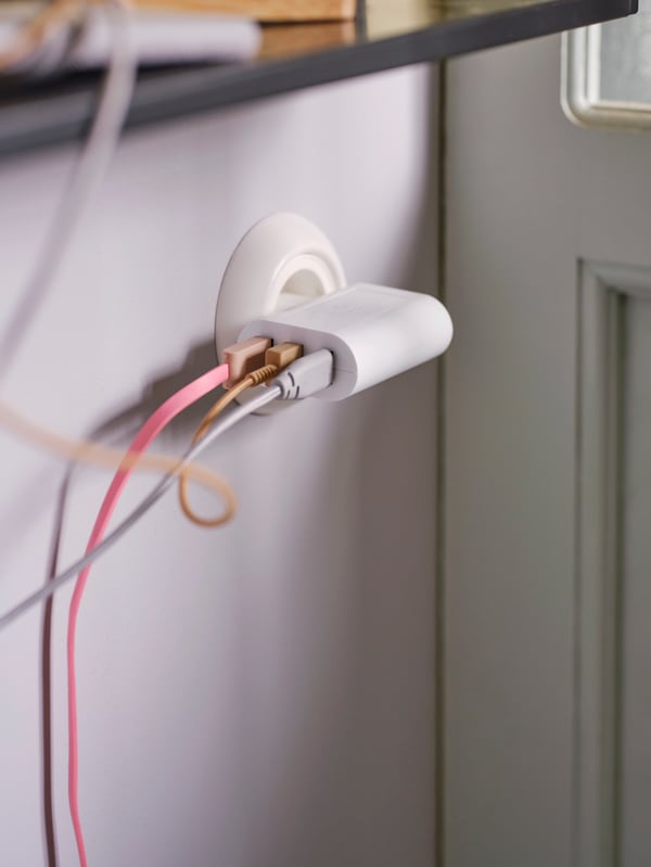 A white SMÅHAGEL 3-port USB charger in a plug socket on a wall with a pink, a yellow and a grey cable in each port.