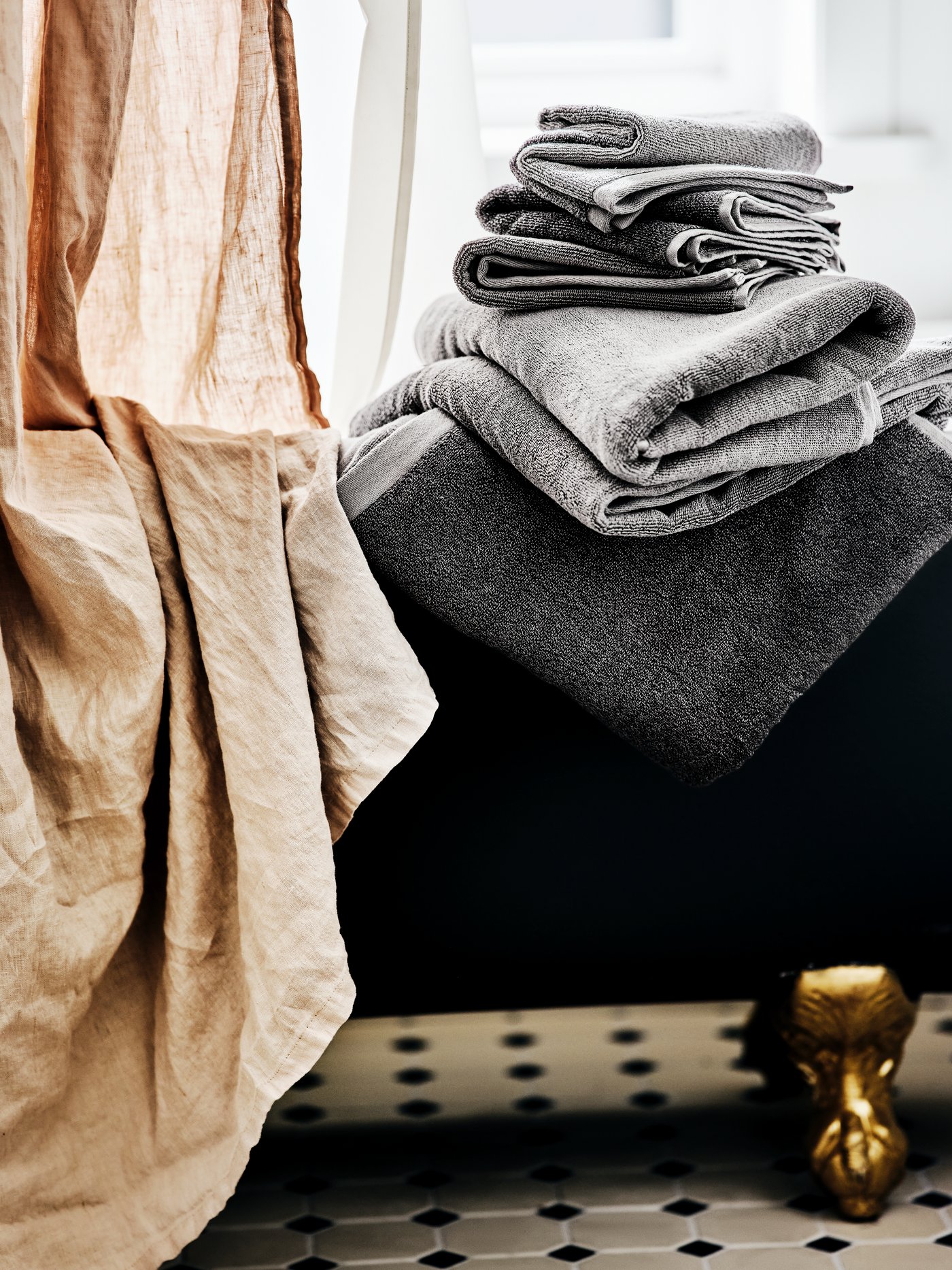 A pile of dark and light grey towels on the side of a black roll-top bath with gold feet, with a beige shower curtain.