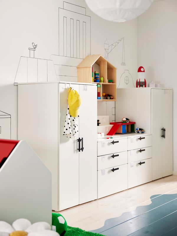 Elementi contenitori SMÅSTAD/PLATSA disposti lungo una parete con dei disegni divertenti a completare la scena, più una casa per bambole FLISAT appoggiata in alto. Sul pavimento è stato dipinto il mare blu.