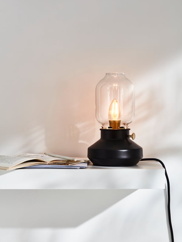 A lamp resembling an old-fashioned candle-stick with a candle, sitting on a white wall shelf beside an open book.