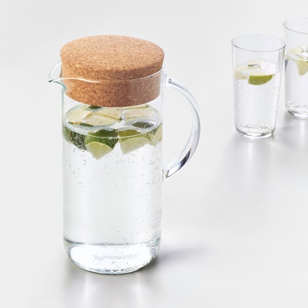Des tranches de citron flottent dans l’eau qui a été versée dans un pichet IKEA 365+ en verre avec un bouchon en liège derrière lequel se trouve un grand verre ; ils sont posés sur une surface blanche. 