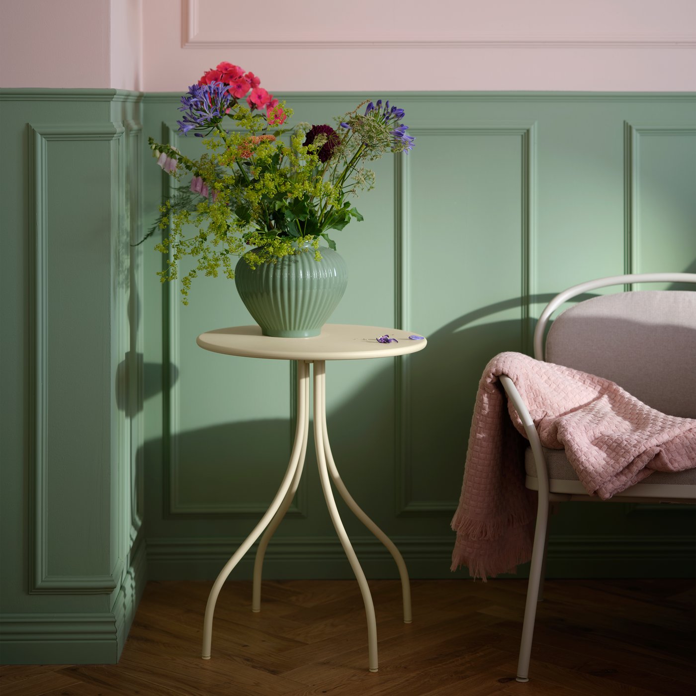 Et lysegult TÅNEBRO sidebord holder en vase med blomster ved siden af ​​en lænestol i et blødt oplyst rum med grønne vægge.