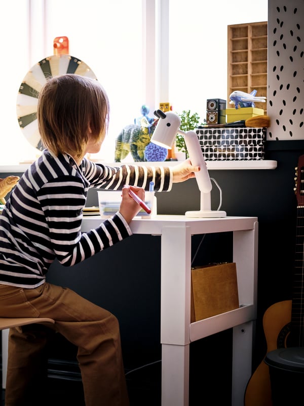 Un niño dibujando sobre un escritorio para niños PÅHL. El niño está prendiendo una lámpra infantil KRUX.