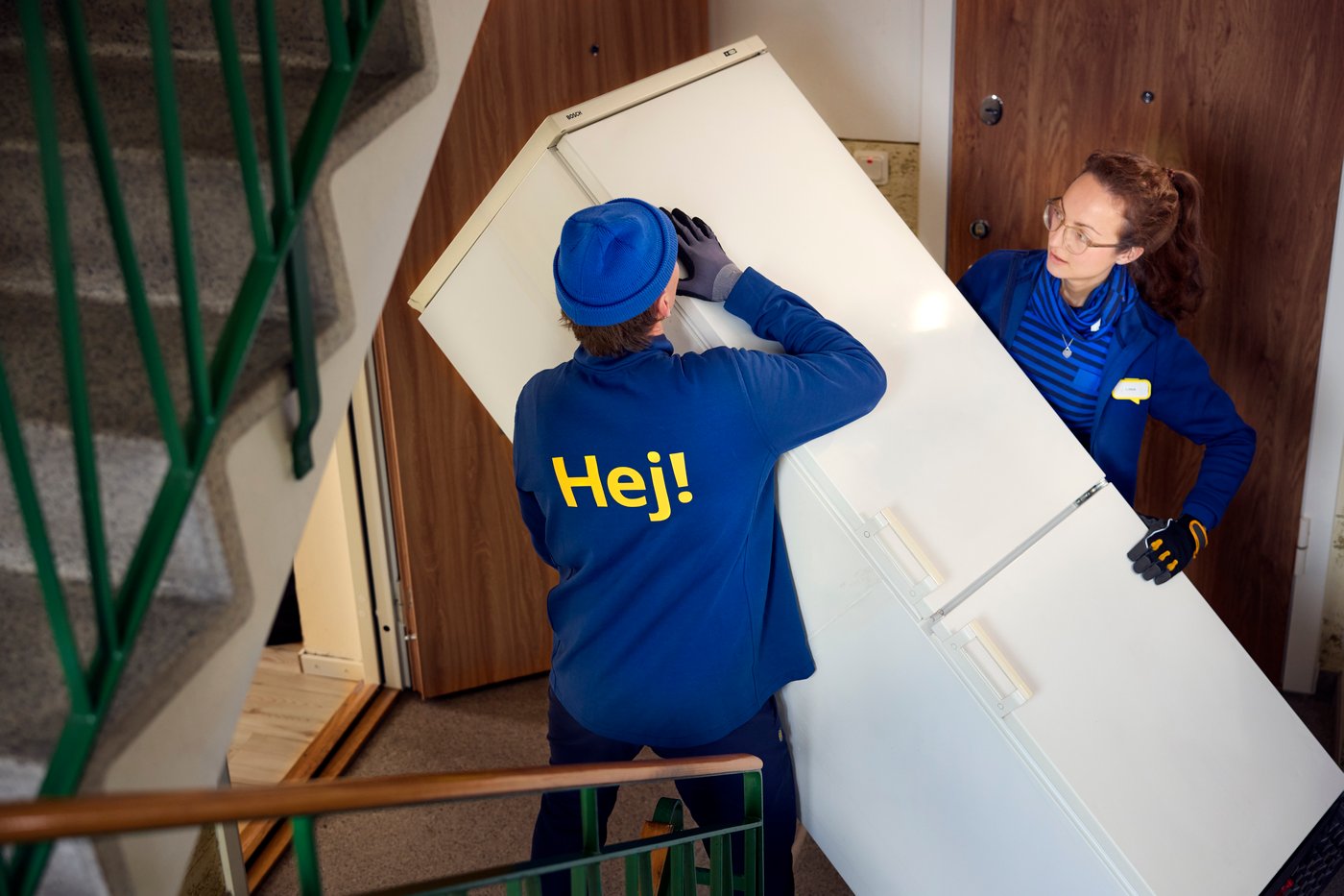 Zwei IKEA Mitarbeiter:innen tragen einen Kühlschrank eine Treppe hinunter.