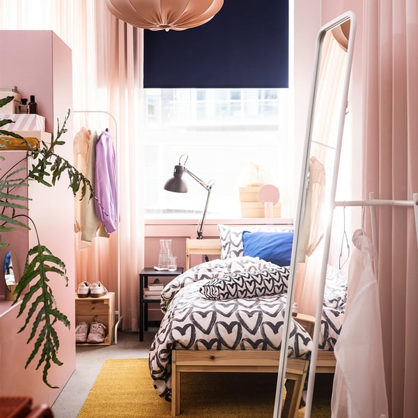 Bedroom with pink vibes, showing a KNAPPER mirror hanging near a wall with clothes hanging behind the mirror. There is a NEIDEN bedframe with black and white tones duvet quilt cover. 
