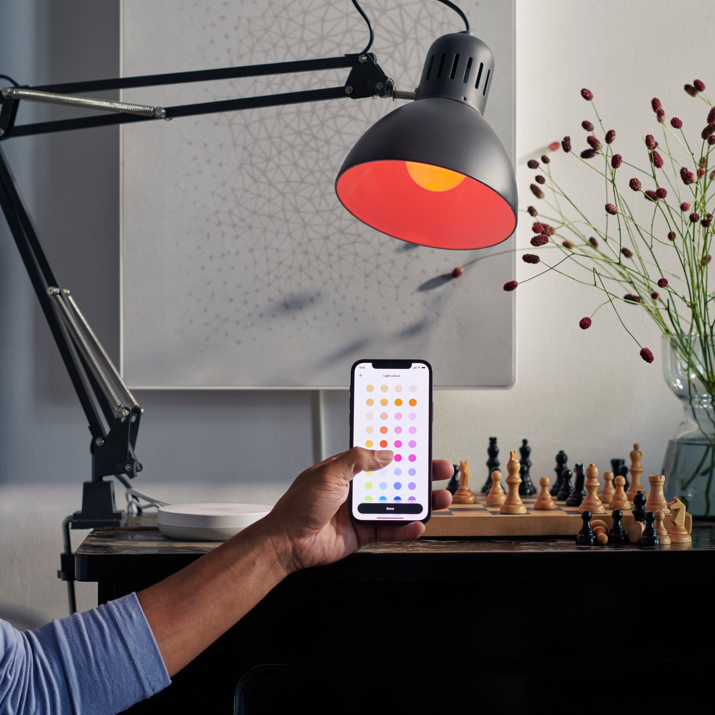Person vid skrivbord med schackbräde, håller smartphone belyst av rödskärmig skrivbordslampa.