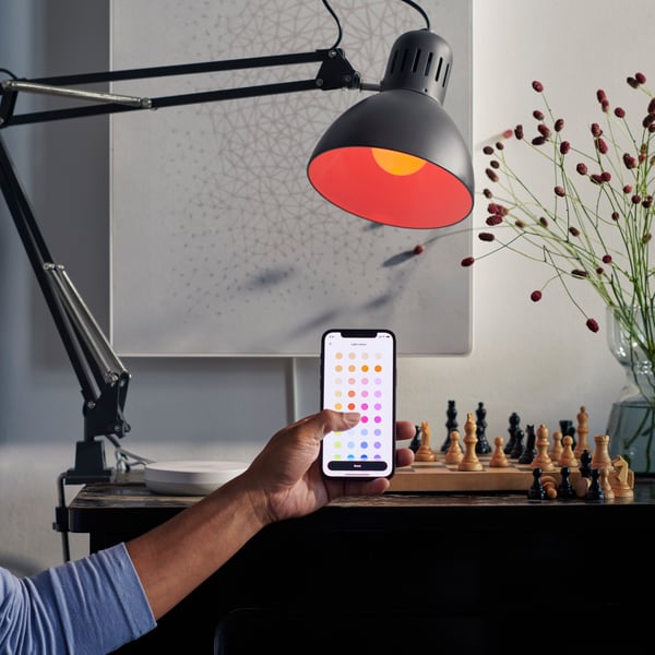Osoba pokazuje aplikaciju IKEA Home smart na pametnom telefonu, šahovska tabla i zvučnik u pozadini.
