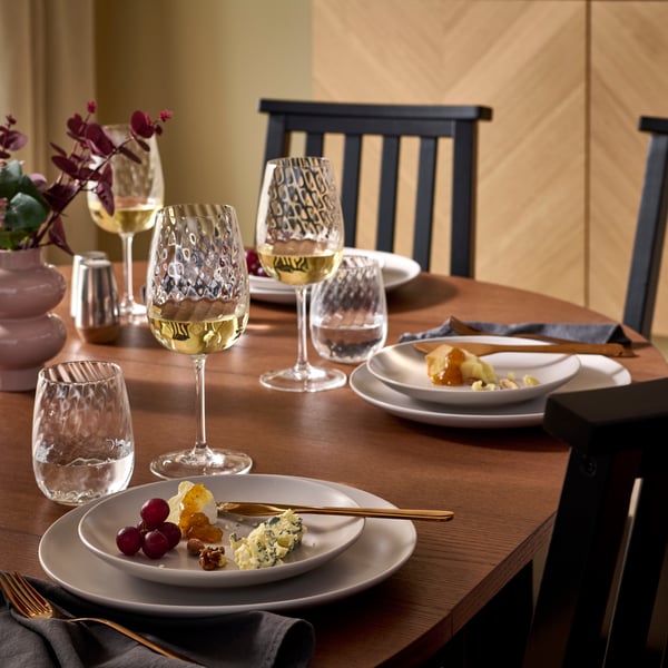 A round ÅLHULT extendable table set for dinner with a matt light grey FÄRGKLAR 18-piece service and FJÄLLUGGLA wine glasses.