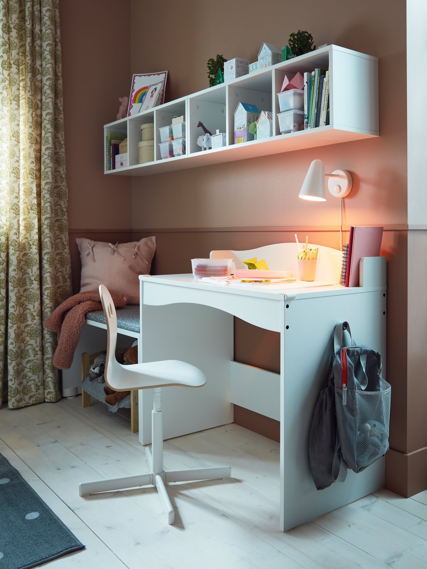 A white desk, a white children's desk chair, a bag, books, a white LED wall lamp, white shelf units.
