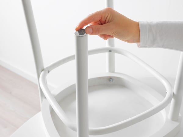 Krupan kadar ruke koja stavlja ili podešava sivi poklopac na gornjem kraju belog, cilindričnog metalnog nogara komada nameštaja, možda stolice ili stola. Pozadina je minimalistička i pretežno bela, što sugeriše unutrašnje okruženje s čistom i jednostavnom estetikom dizajna.