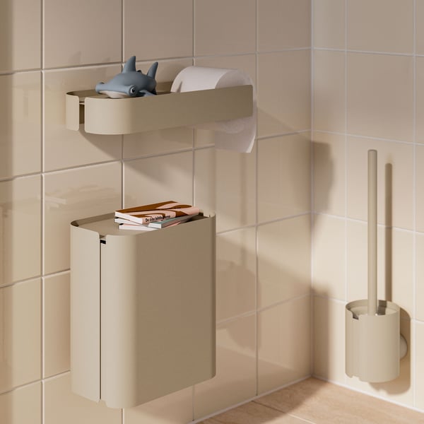 A tiled bathroom corner by a toilet has a grey-beige FLORSJÖN waste bin mounted on the wall, with books on its recessed lid.