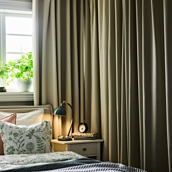 A bedroom with BENGTA block-out curtains next to a neatly made bed, bedside table with a lamp and alarm clock.