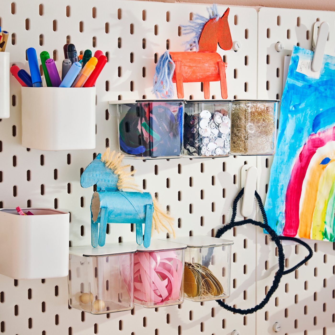 A SKÅDIS pegboard in white on a wall, with containers and cups holding pens, craft supplies and small toys attached to it.