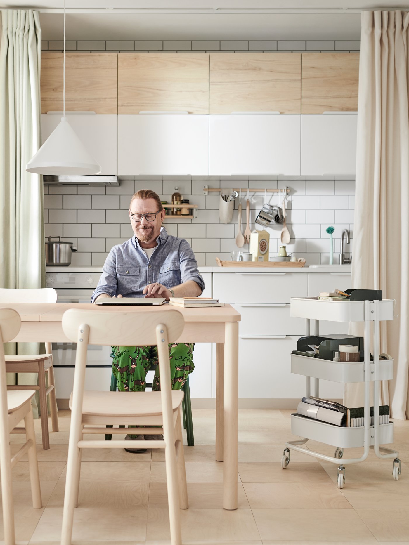 A man opens his computer on a RÖNNINGE extendable table in a kitchen with white VEDDINGE and ash effect ASKERSUND doors.