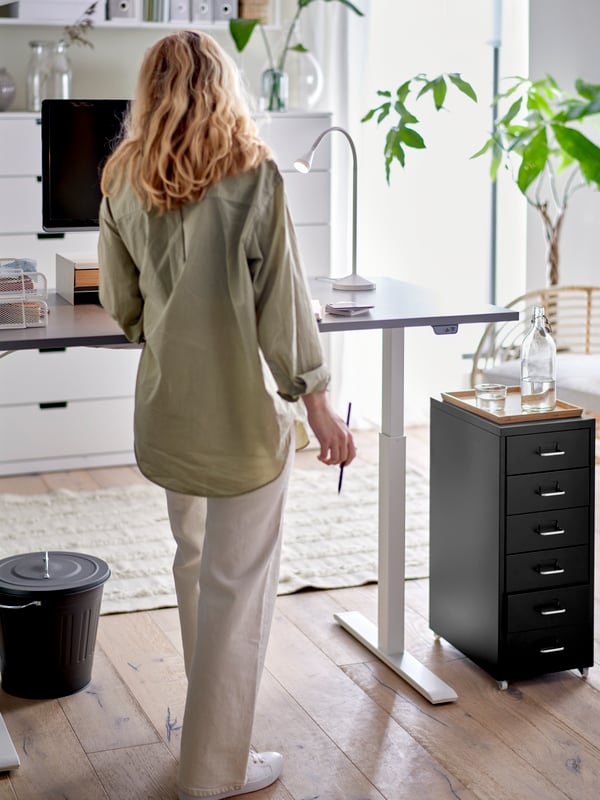A home office with a woman standing at a grey sit/stand RODULF desk with a computer, a work lamp, and a drawer unit.