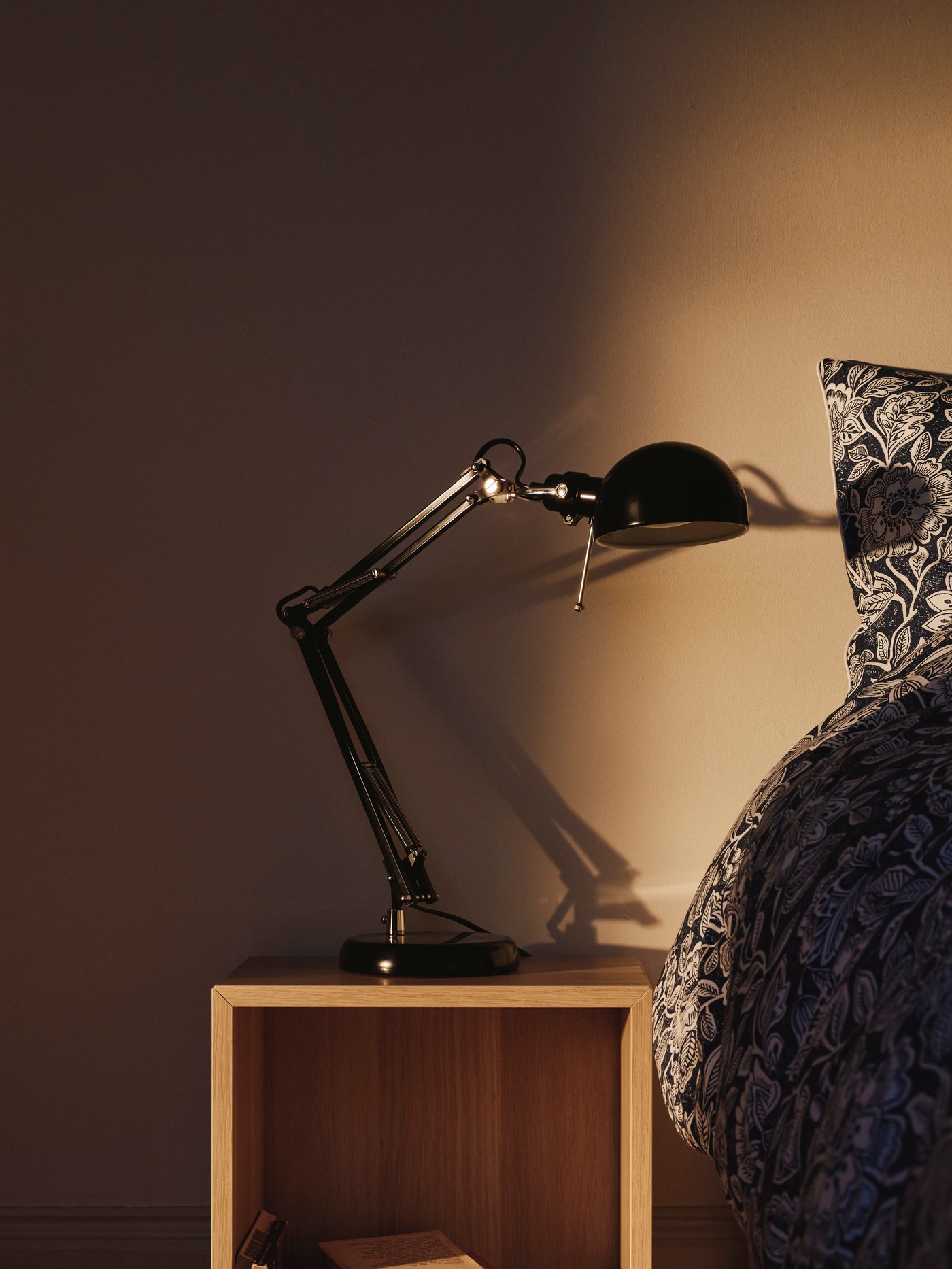 En svart justerbar skrivbordslampa står på ett öppet sängbord bredvid en säng med mörkt mönstrat täcke, upplyst av varmt ljus.