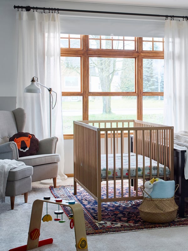Corner of a bedroom with a wooden baby’s cot. An armchair on one side, a baby gym on another, a mobile hanging above.