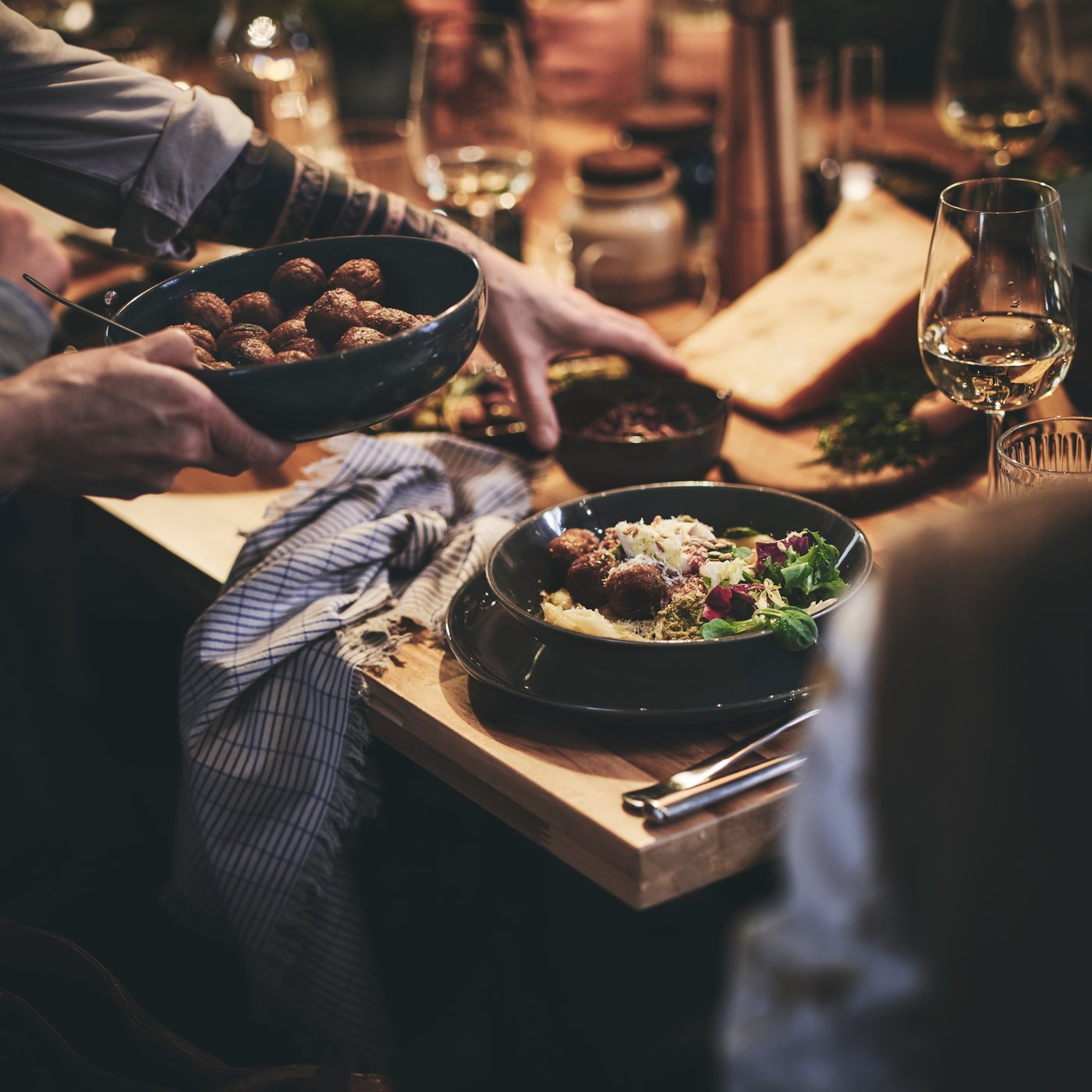 A dinner guest at a long table passes a bowl of HUVUDROLL plant balls to the guest sitting beside them.