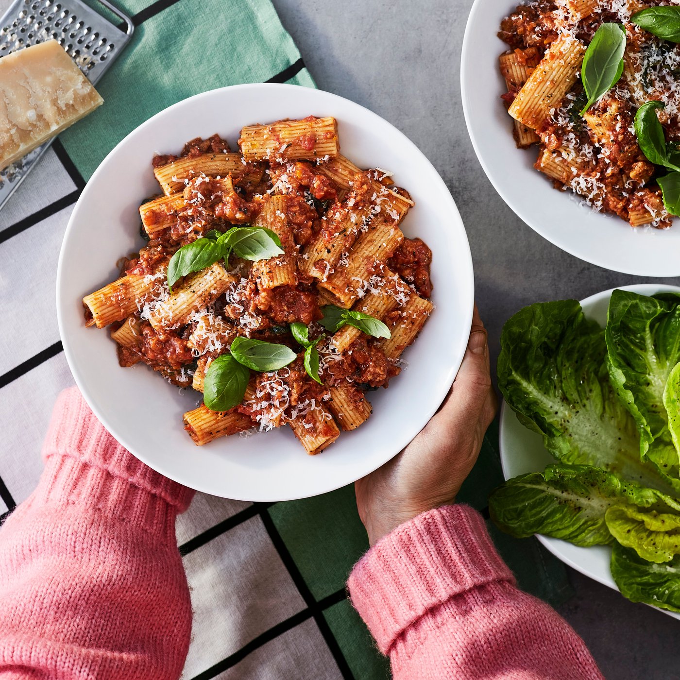 En person iført en lyserød sweater holder en tallerken med plantebaseret bolognese og pasta med en smule basilikum på toppen.