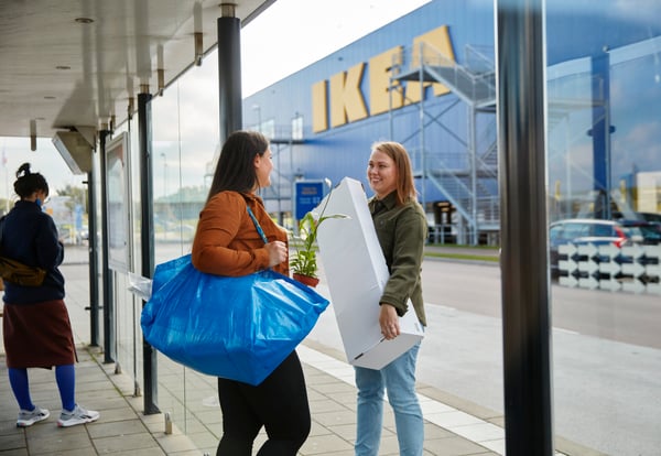 To kvinder snakker sammen foran et IKEA varehus, den ene bærer en blå FRAKTA bærepose over skulderen, mens den anden bærer en hvid pakke.