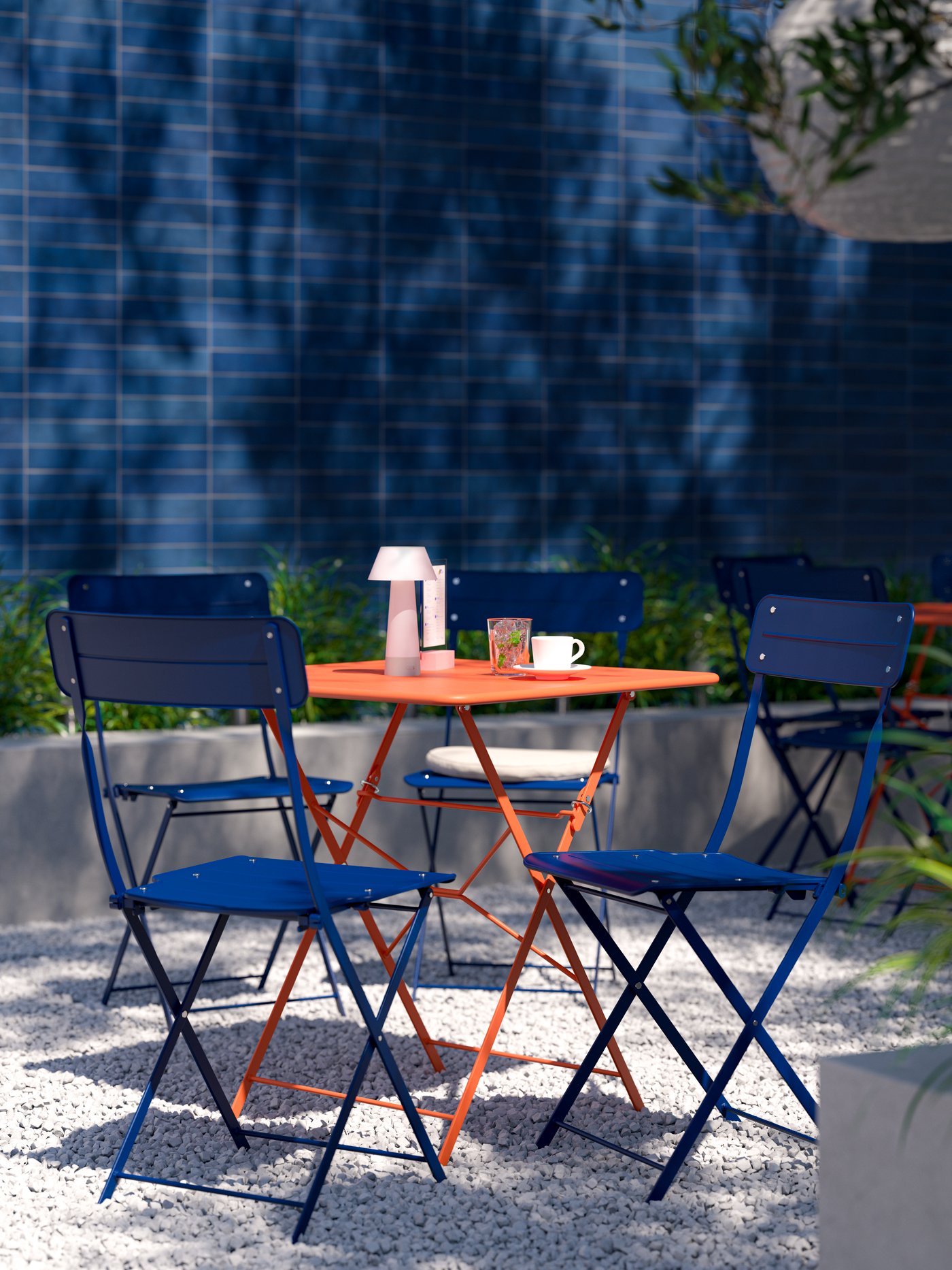 Four dark blue SUNDSÖ outdoor chairs and a bright orange SUNDSÖ outdoor table with a table lamp in a sunny outdoor space.