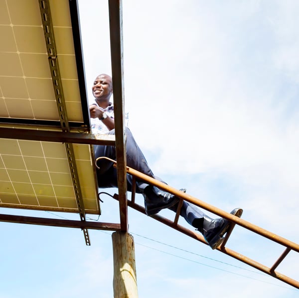 A person atop a ladder inspects the top side of a solar panel on a bright sunny day.